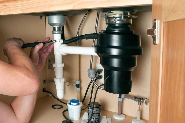 Plumber repairing a garbage disposal beneath a kitchen sink in Holladay